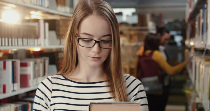 Close up of young female student standing in university library pulls book off shelf and begins to study preparing for lectures.