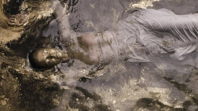 A woman submerged in water, covered all over with gold paint. He lies with his eyes closed and runs his hands through the water.