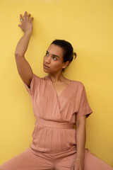 Obraz premium female model woman sitting with raised arm, wearing a pink casual jumpsuit and hair collected, studio with yellow background