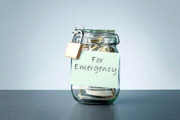 Money glass jar with written sticky note FOR EMERGENCY on gray background Concept of setting money...