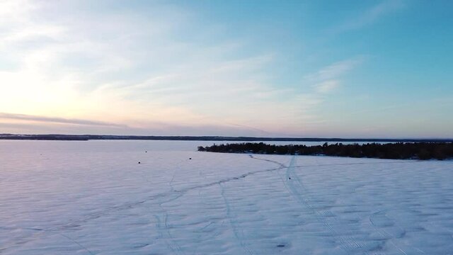Frozen snow covered lake road with ice fishing huts, ice road and sled during sunset; wide aerial