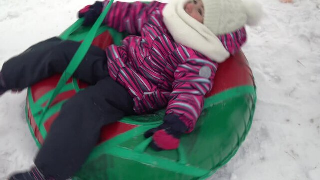 The girl is being spun on an inflatable ring in the snow. A girl in warm clothes lies on an inflatable ring and is unwound with a rope