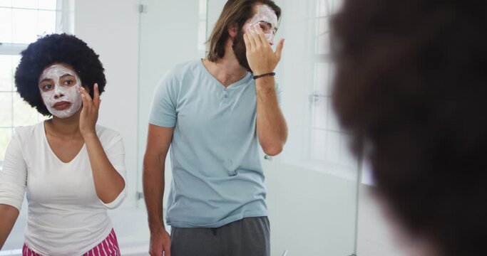 Mixed race couple applying face mask together while looking in the mirror at bathroom