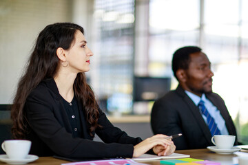 Attractive young adult caucasian businesswoman sitting and concentrated listening to presentation in a multiracial business meeting. Selective focus at woman face with a blurred African businessman