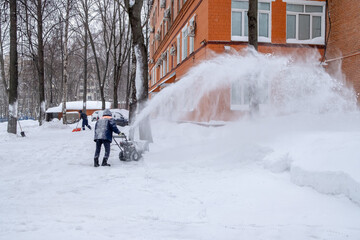 A utility worker cleans a path with a snow blower during heavy snow on a winter day. A jet of snow takes off into the air.