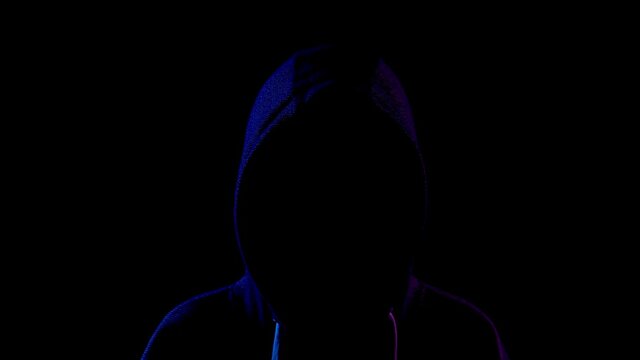 A young woman is standing in the hood hiding her face. An attacker is standing in the dark. Blue and red light falls on the person on a black background.