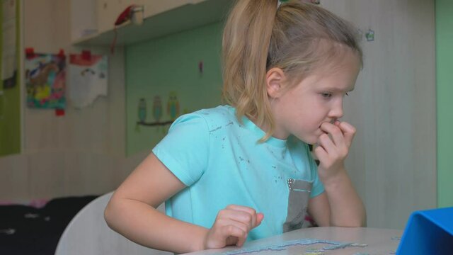 In the children's room with drawings on the wall, a girl with blond straight hair, a ponytail hairstyle, holds a finger in her mouth and looks into a smartphone, worries.