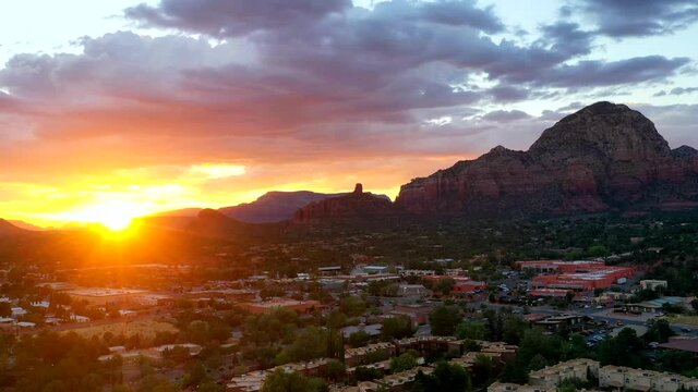 A hyperlapse of the sun setting over Sedona as traffic bustles. The sun peaks through the clouds radiating the sky with bursting colors of red, pink and orange with towering red rocks in the picture.