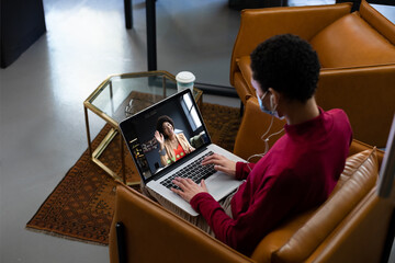 African american woman wearing face mask having video call with female colleague displayed on laptop