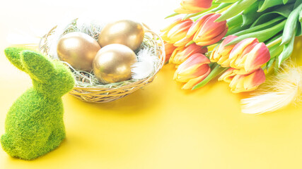 Golden eggs in basket with spring tulips, white feathers on pastel yellow background in Happy Easter decoration. Foil minimalist egg design, modern design.