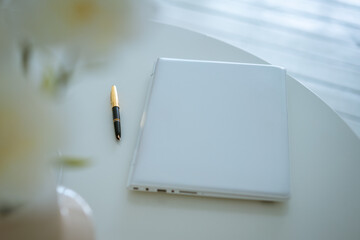 Notebook and pen on a white table, flowers in a vase. Soft focus