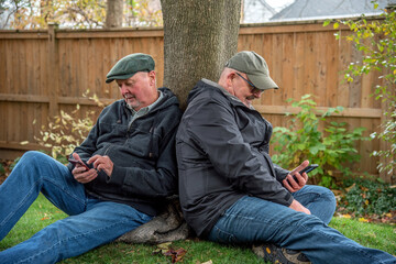 LGBT gay senior married couple sitting against a tree and checking their mobile phones.