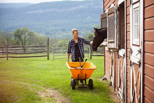 Rancher stroking horse while pushing wheelbarrow in farm
