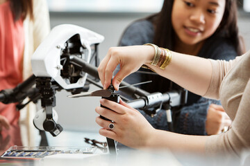 Female student fixing propeller on drone with teacher and friend in background