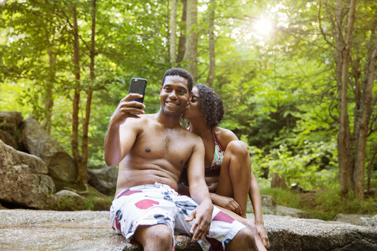 Woman kissing man taking selfie through smart phone on river bank in forest