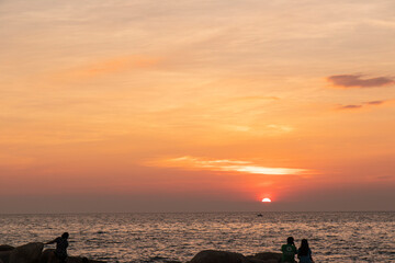 The sun on beach in evening have the background of the sea in the evening. There are people standing and site on rocks