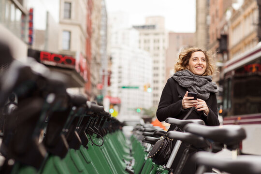 Woman holding mobile phone standing by bicycle rack in city