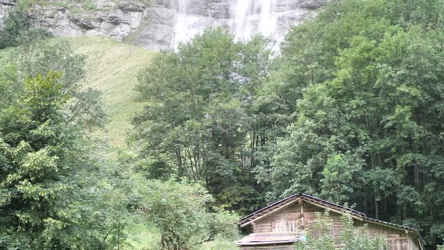 Waterfall falling down on the background behind trees with a lovely wooden challet on the foreground in Lauterbrunnen Switserland. Wide shot