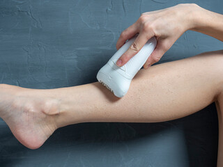 a woman shaves her leg with an electric epilator on a gray textured background. The concept of beauty and self-care