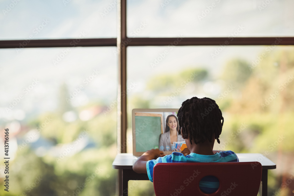 Poster Rear view of male student having a video call with female teacher on laptop at school