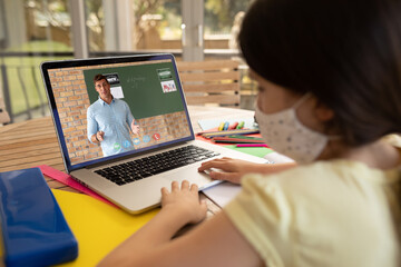 Female student wearing face mask having a video call with male teacher on laptop at school