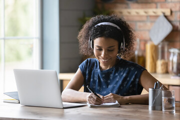 Close up smiling African American woman in headphones writing taking notes, using laptop, listening...