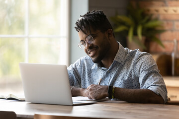 Close up smiling African American man wearing glasses using laptop, writing, taking notes, student watching webinar, studying online, looking at computer screen, sitting at wooden desk at home