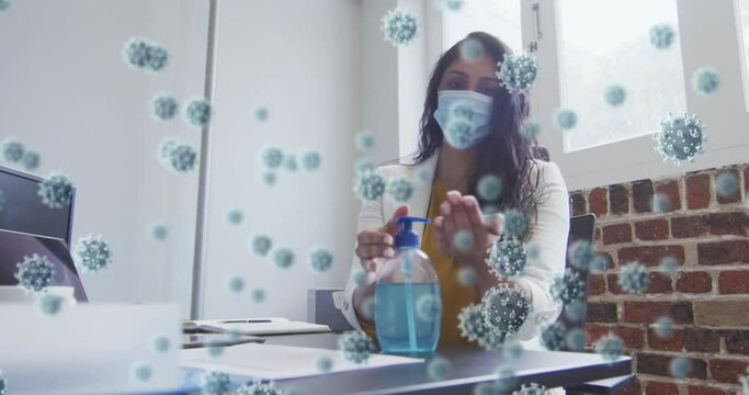 Animation of covid 19 cells floating over mixed race woman wearing face mask disinfecting her hands 