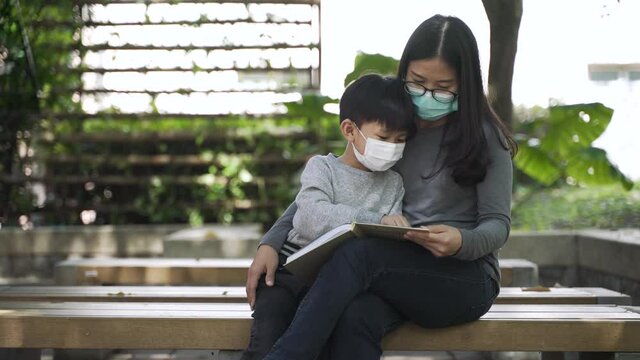 Asian boy about 5 years old with face mask reading the book with his mother at the public garden park