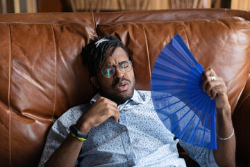 Close up exhausted African American man wearing glasses waving paper fan, cooling, suffering from...