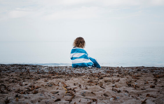 Rear view of girl wrapped in towel sitting at lakeshore against sky