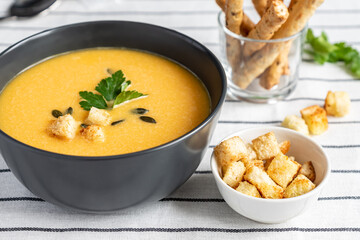 A bowl of homemade fresh spicy organic creamy pumpkin soup on a white striped textile background with bread sticks, sour cream, parsley and spices. Winter and autumn vegetarian food concept.