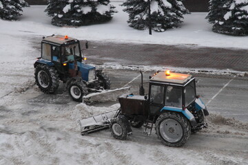 Two wheeled traktor machines cleaning a snow with scraper shovel blade on supermarket parking place after heavy snowfall at winter evening, top side view