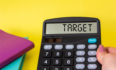 A female hand holds a calculator with the text on the display TARGET on a yellow background, next to colored notepads