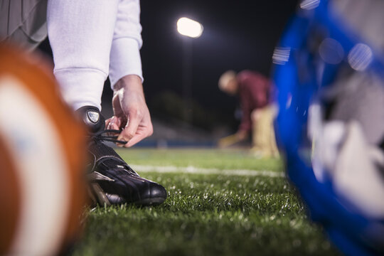 Low section of American football player tying shoelace on field with coach in background