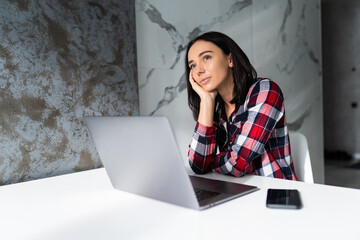 Beautiful sad young woman at the kitchen indoors at home using laptop.