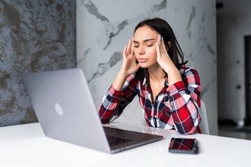 Exhausted tired woman working using on laptop holding head in hand sitting in kitchen at home. Young woman studying or working indoors. Freelance business quarantine concept