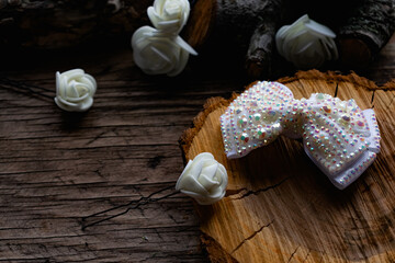 Jewelry and hair accessories. Elegant bow with holographic stones and rhinestones with a pink tint. There are cute milky roses around. Costume jewelry on an old wooden background covering