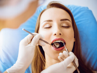 Smiling young woman with orthodontic brackets examined by dentist in sunny dental clinic. Healthy teeth and medicine concept