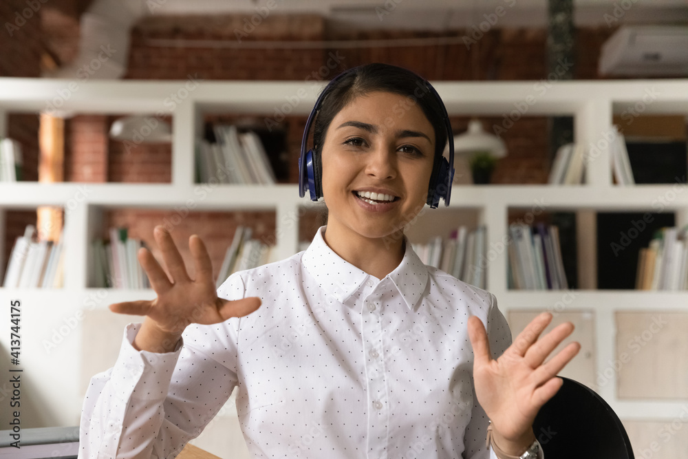 Canvas Prints Portrait of smiling young Indian female employee in headphones talk speak on video call with client or customer. Happy millennial ethnic woman have webcam virtual online call or event in office.