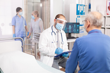 Obraz premium Doctor wearing visor as safety precaution in the course of consultation of senior man. Physician practitioner in the course of private modern clinic appointment during coronavirus pandemic.