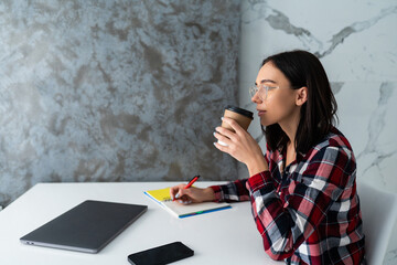 Young pretty woman is sitting at the table in her kitchen and writing in a notebook.