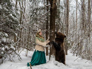 Russian beauty in folk national dress collects snowdrops in the winter forest with a brown bear