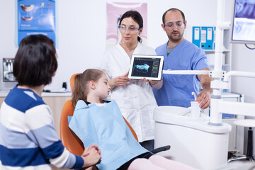 Obraz premium Dentist in dentistiry office showing tablet pc with jaw radiography explaining cavity treatment. Stomatologist explaining teeth diagnosis to mother of child in health clinic holding x-ray.