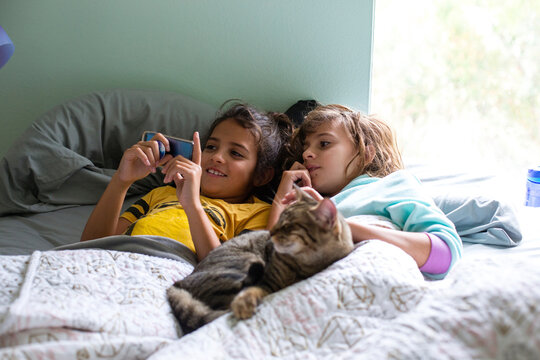 Sisters looking at smart phone while lying with cat on bed