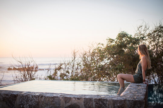 Side view of woman sitting on retaining wall at artificial pond against sea and clear sky