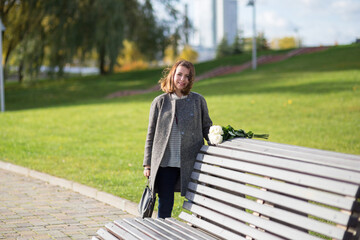 A woman in a coat stands next to a bench in a sunny autumn park. There is a bouquet of white roses on the bench. The woman is smiling, her hair is tousled by the wind.