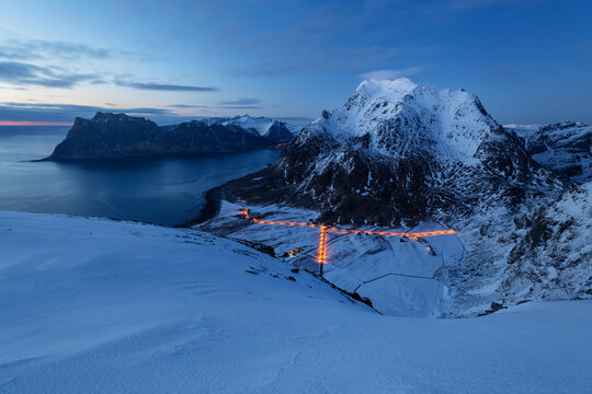 Streelights of Uttakleiv village shine below Himmeltindan mountain peaks in winter twilight, Vestvågøy, Lofoten Islands, Norway