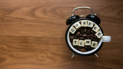 Top view a cup of coffee beans on top of alarm clock with alphabet of text Coffee Time on blurry wooden background with copy space.