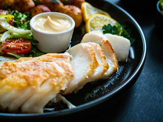 Fish dish - fried cod fillet with potatoes and vegetable salad on black wooden table